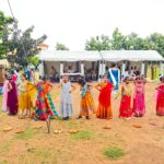 Group of young girls in colorful traditional indian outfits lined up outdoors at a festive event with white tents in the background